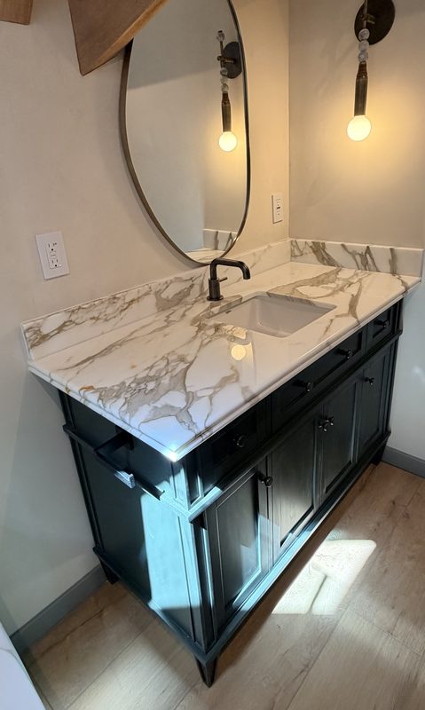 Dark green bathroom vanity with a Calacatta Gold marble top, round mirror and brass sconce